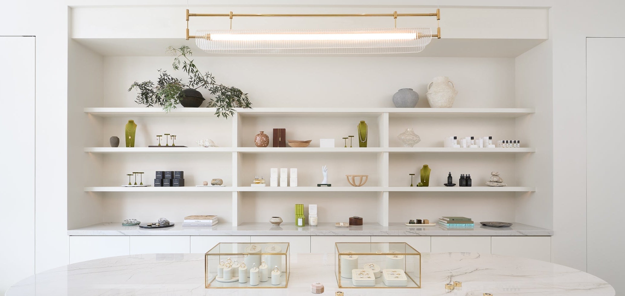 A modern interior scene featuring a large oval marble-top table with a fluted wood base, set in front of built-in white shelves with decorative objects. The room has a bright, airy aesthetic with white walls, recessed lighting, and a gold linear light fixture.
