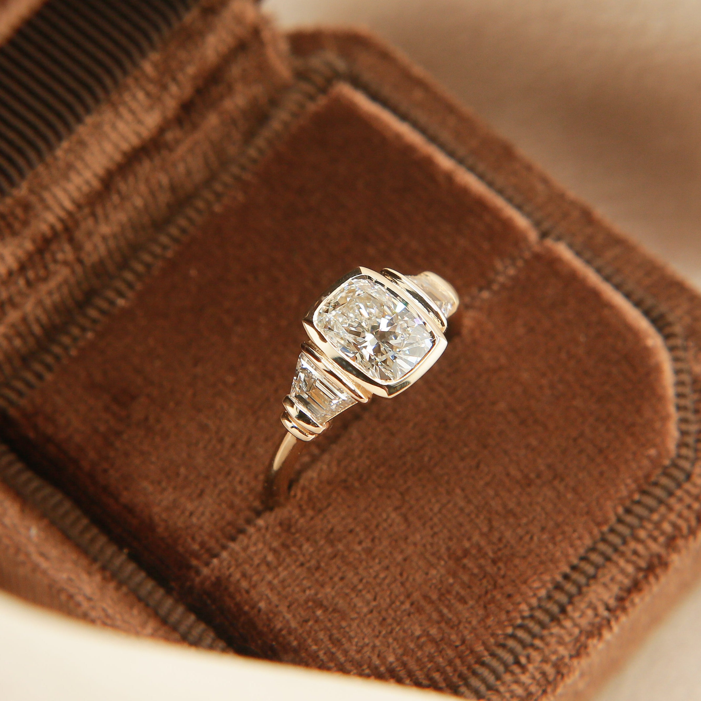 Cushion cut diamond three stone engagement, tab set trapezoid side stones in a velvet box.