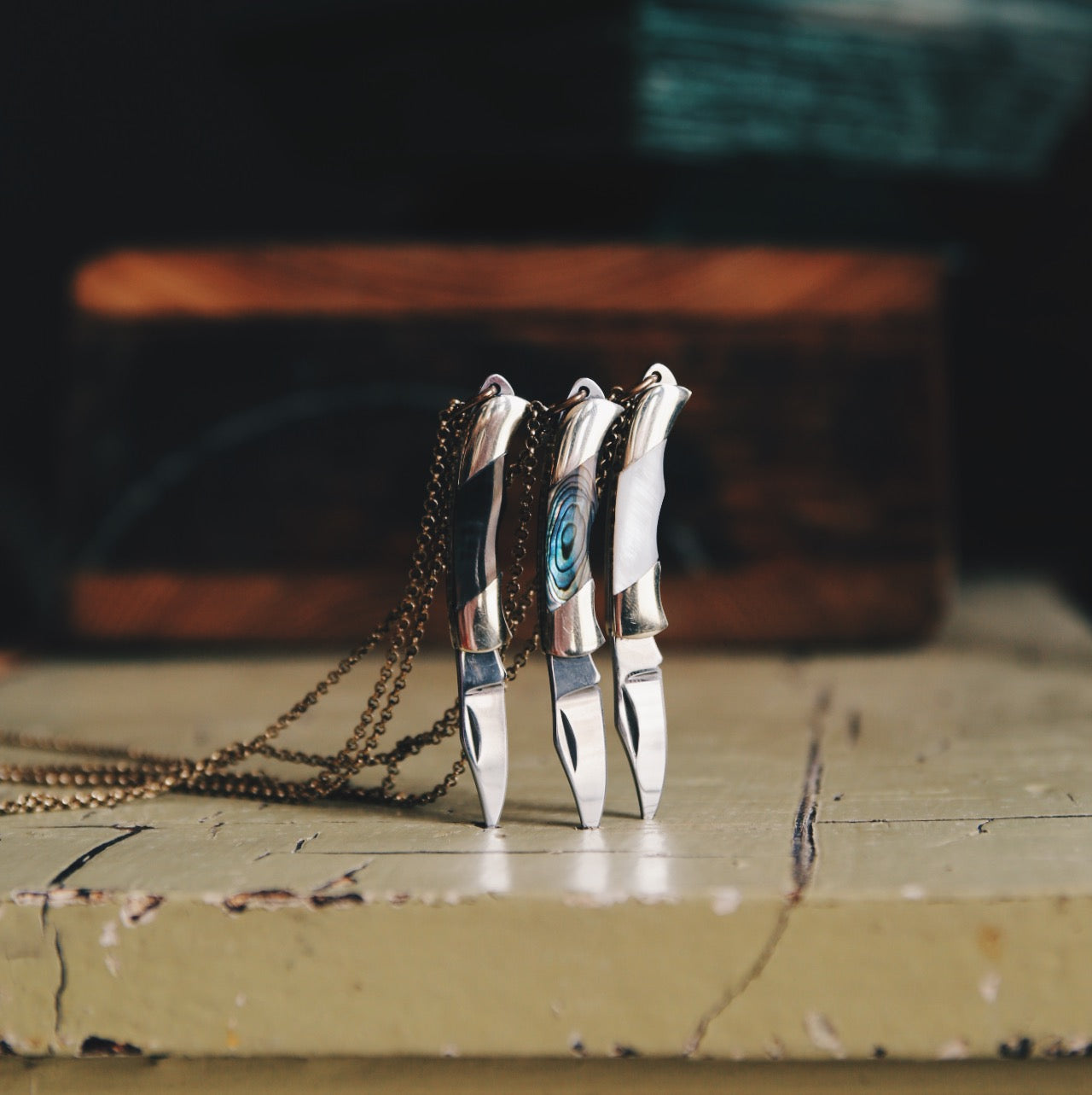 Three pocket knife necklaces with blades slightly sunk into a table so they're standing upright. From Cival Collective, a Jewelry Store in Milwaukee Wisconsin