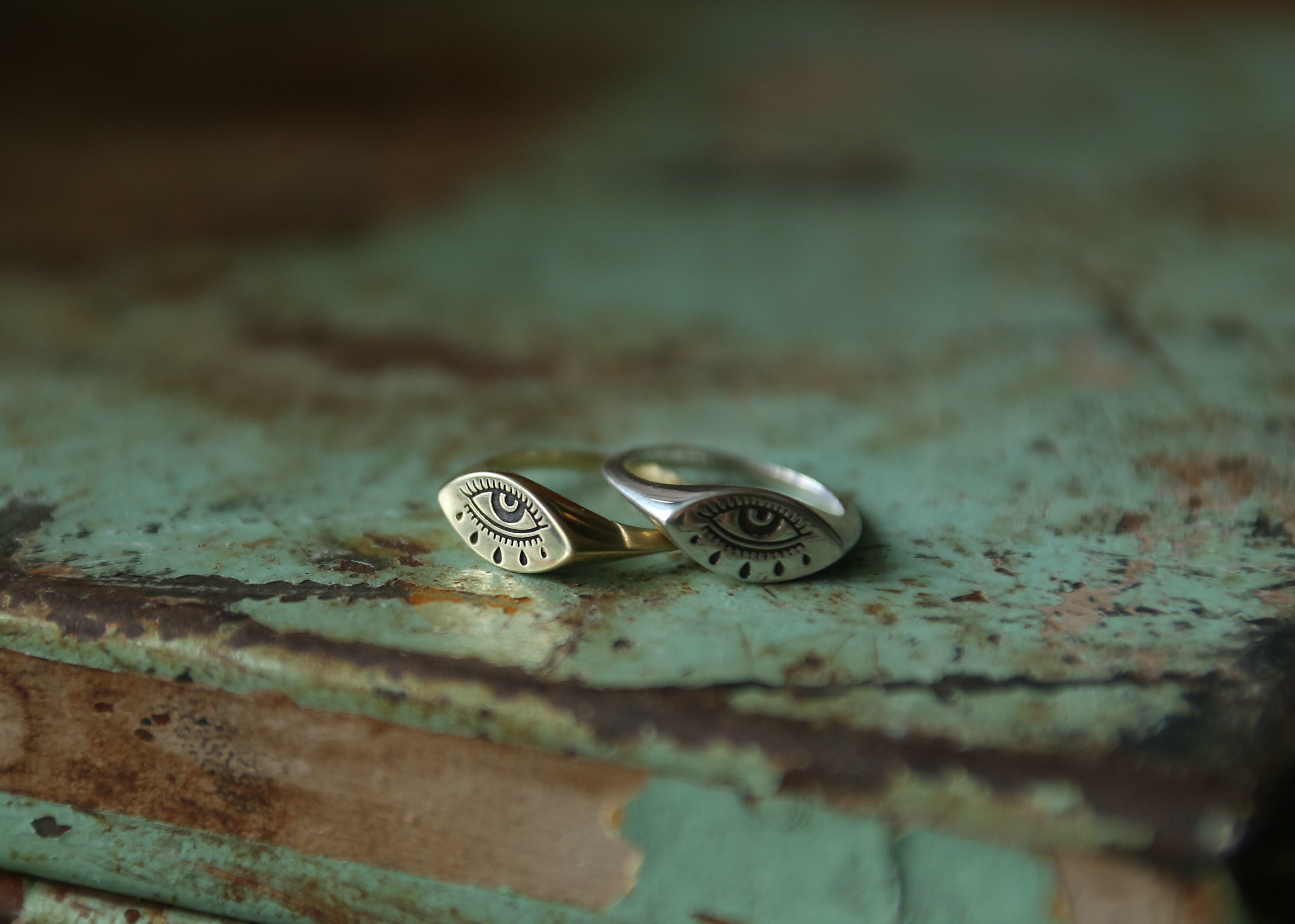 brass and silver evil eye rings next to each other slightly overlapping, on blue-green background