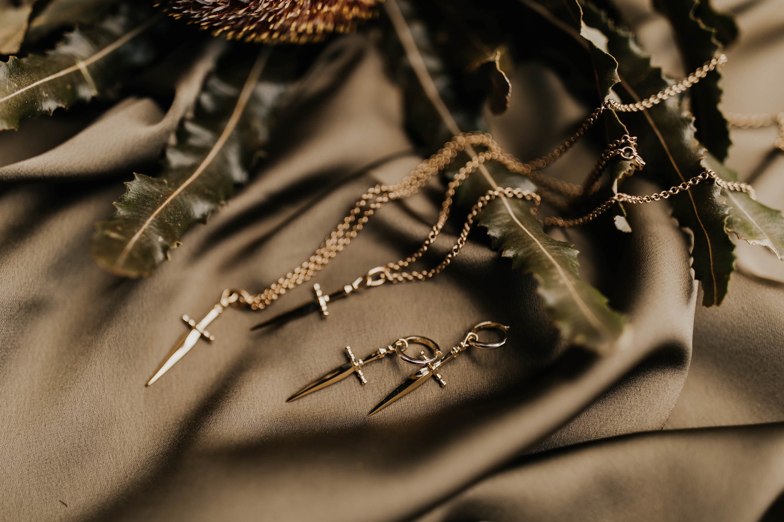 brass hoop earrings with swords dangling down laid on table, necklaces laid next to them also with sword pendant 